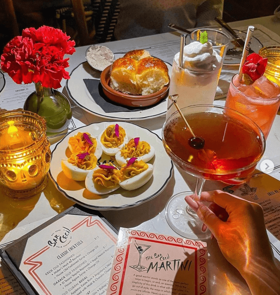 A table is set with cocktails, deviled eggs, a bread roll, flowers, and illuminated candles, alongside a menu.