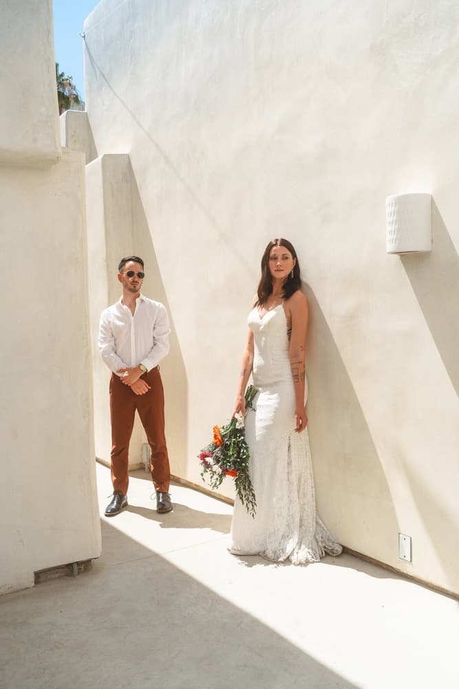 A couple stands in a sunlit corridor, with the woman in a white wedding dress holding a bouquet and the man in a white shirt and brown pants nearby.