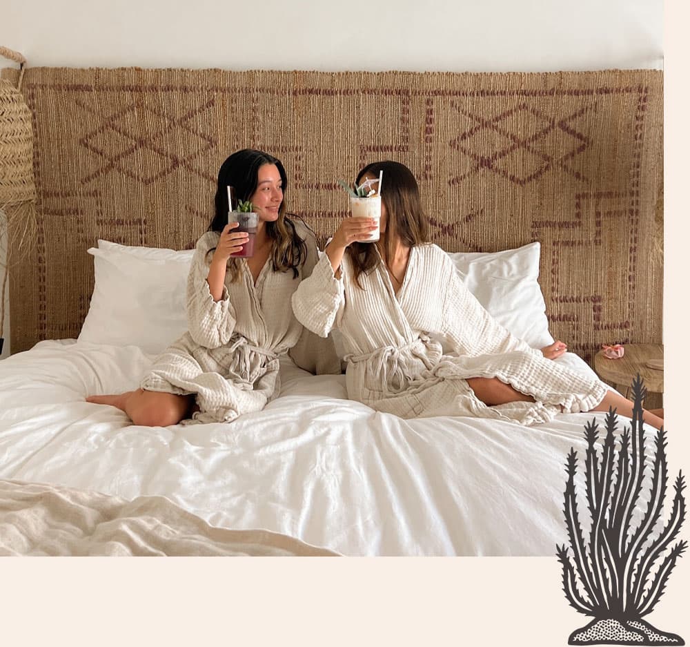 Two women in bathrobes sit on a bed, raising their drinks in a celebratory toast.