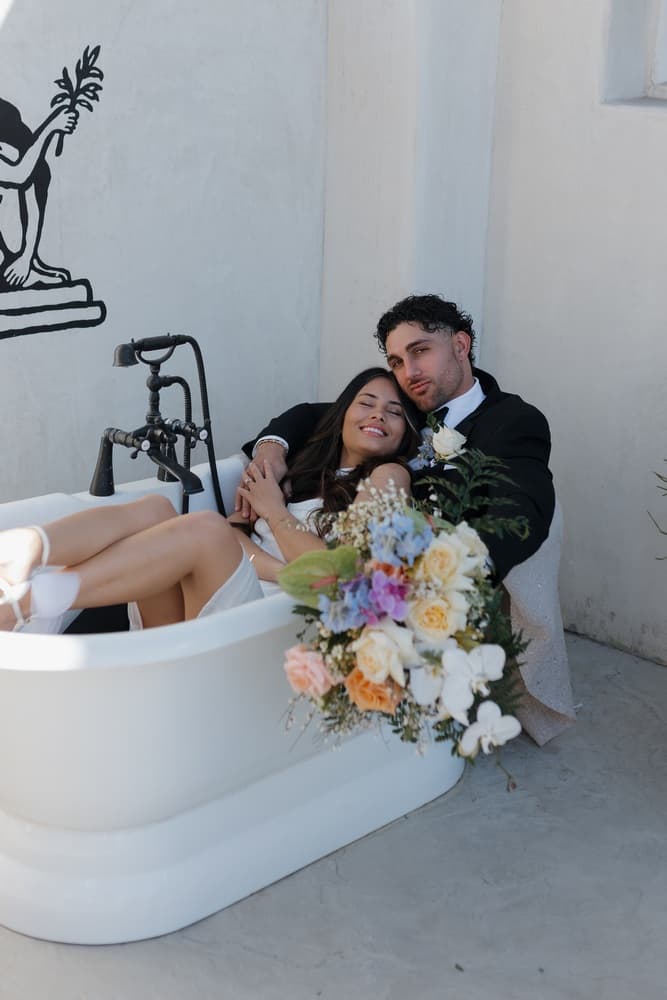 A couple relaxes in a white bathtub holding a colorful bouquet, surrounded by a minimalist backdrop.