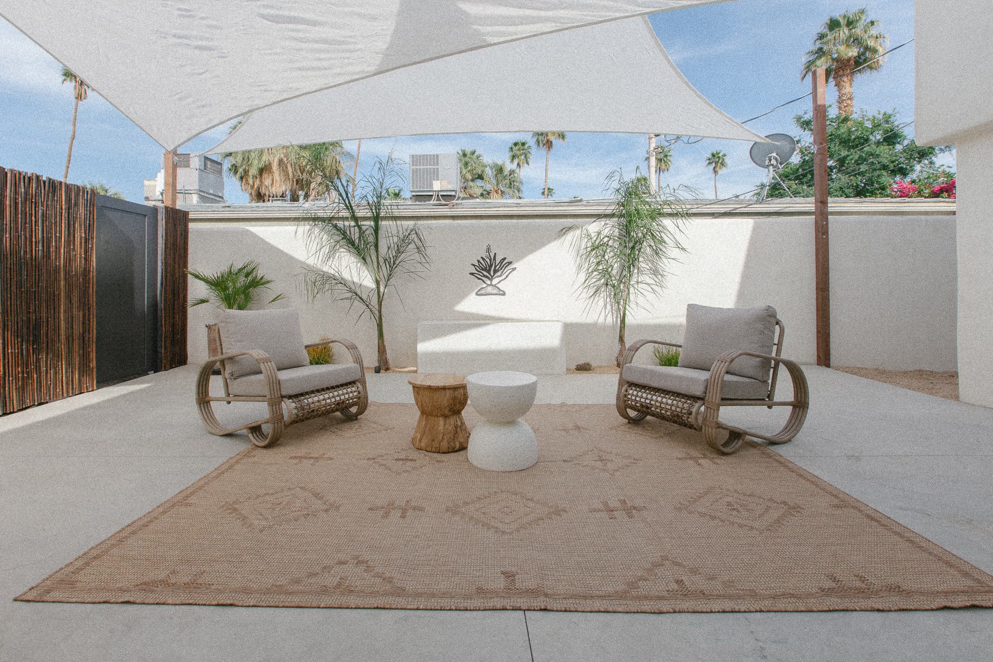 A private, sunny patio area featuring two curved wooden armchairs with cushions, a geometric patterned rug, natural fiber side tables, and a white sun shade canopy overhead.
