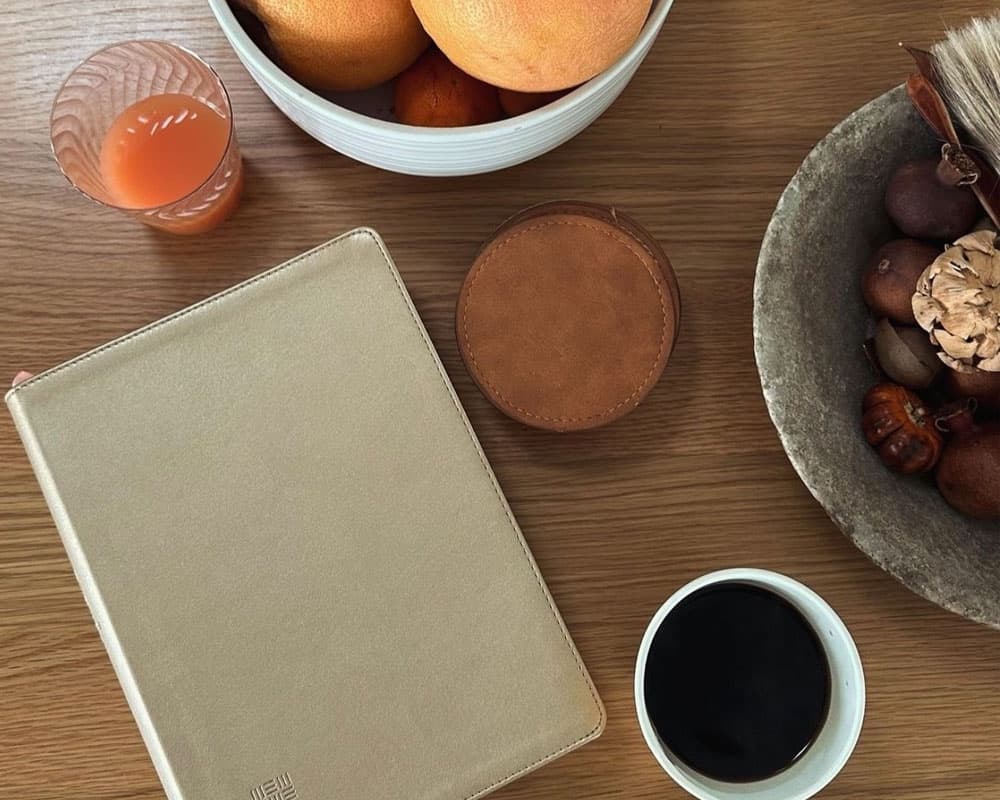 A top-down view of a wooden table featuring a bowl of oranges, a glass of juice, a notebook, a leather coaster, and a cup of coffee.