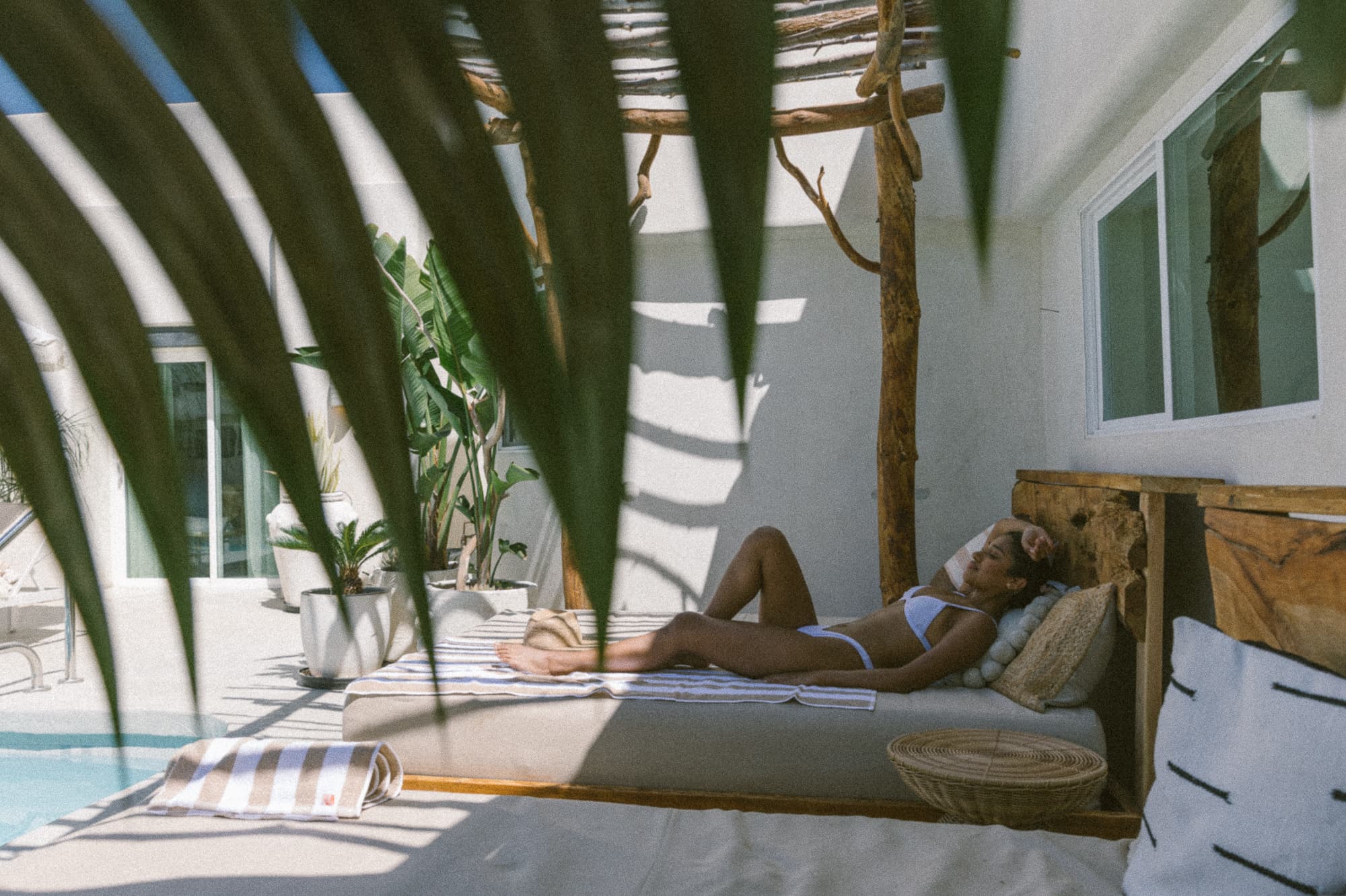 A woman lounges by a pool on a sunny day, framed by tropical greenery.