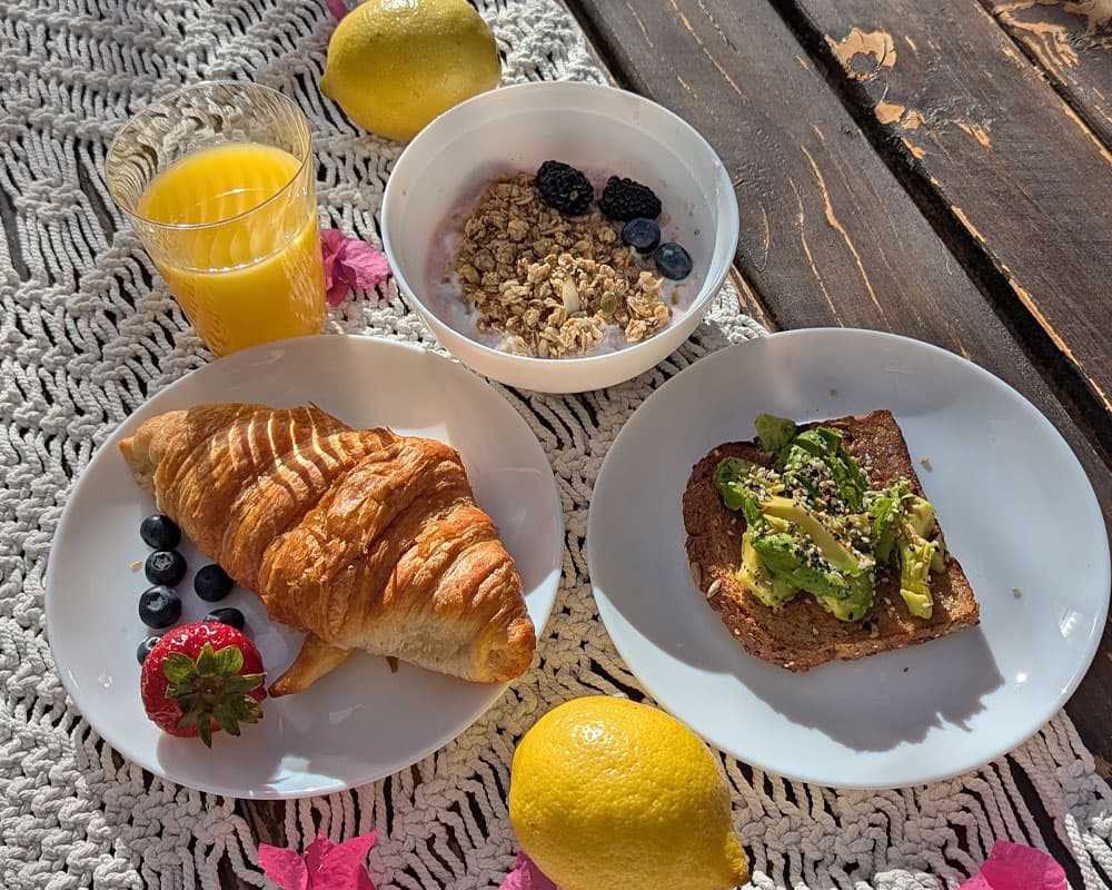 A breakfast spread featuring a croissant, toast with avocado, a bowl of granola with berries, and a glass of orange juice, surrounded by lemons and pink leaves on a lace tablecloth.