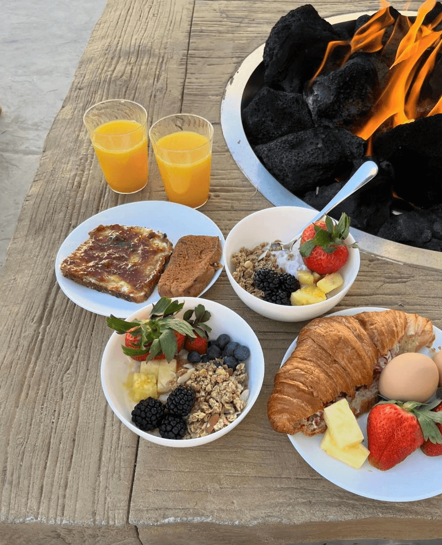 A variety of breakfast dishes including fruit, yogurt, croissant, and orange drinks are arranged on a wooden table near a fire pit.