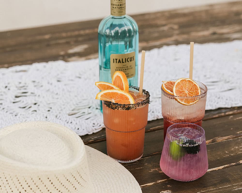 Three colorful cocktails garnished with orange slices and a bottle of Italicus, beside a straw hat on a wooden table.
